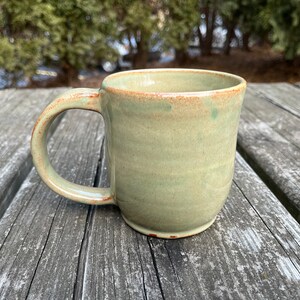 May include: A light green ceramic mug with a brown rim and handle. The mug is sitting on a wooden surface.