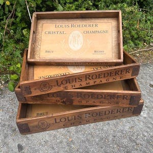 May include: A stack of three rectangular wooden trays with dark brown stained sides and light tan interior bases. Each tray features the black printed text LOUIS ROEDERER CHAMPAGNE, the word BRUT, and a circular logo with the letters LR in the centre.