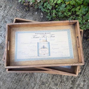 May include: A rectangular wooden serving tray with a light brown finish and cutout handles. The centre features a decorative print with the text "Fortnum & Mason Time For Tea" and an illustration of a tea jar surrounded by various tea names.