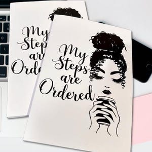 Op de afbeelding: Twee witte notitieboekjes met de tekst "My Steps are Ordered" in elegant schrift. Eén notitieboekje heeft een zwart-wit illustratie van een vrouw met een koffiekopje. De notitieboekjes liggen op een wit oppervlak.
