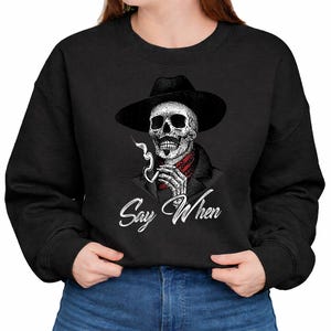 May include: Black sweatshirt featuring a detailed skull illustration wearing a black hat and red bandana, smoking a cigarette. The phrase "Say When" is written in white script below the skull. A unique graphic design.