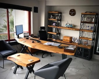 May include: A large wooden desk with a live edge top and black metal legs. The desk is in a modern office with a large window, two black chairs, and a wooden shelf unit with black metal accents.