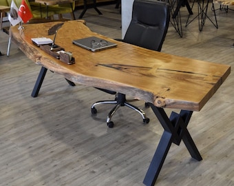 May include: A large, natural wood desk with a live edge top and black metal X-shaped legs. The desk has a laptop, glasses, and office supplies on the surface.