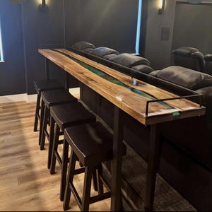 Peut inclure: Un long bar en bois avec un cadre en métal noir et une rivière d'époxy verte traversant le centre. Le bar est installé dans un home cinéma et comporte quatre tabourets de bar noirs.