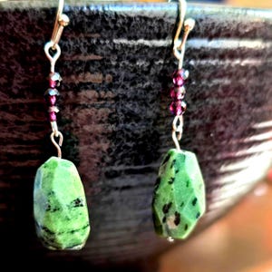May include: A pair of dangle earrings featuring faceted green stones with black inclusions. The earrings have silver-colored hooks and are accented with small, dark red beads. The earrings are displayed against a dark, textured background.