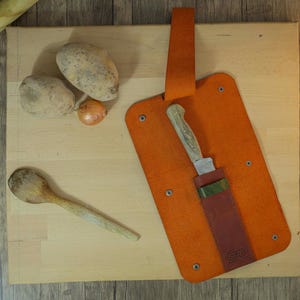 Puede incluir: Vista aérea de una tabla de cortar de madera con patatas, una cebolla y una cuchara de madera. Una funda de cuero naranja para cuchillos con un cuchillo está en la tabla. Un rodillo está en la esquina superior izquierda.