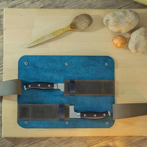 May include: A blue leather knife roll with two knives, a wooden spoon, potatoes, and an onion on a wooden cutting board. The knives have black handles and silver blades. The cutting board is light brown.