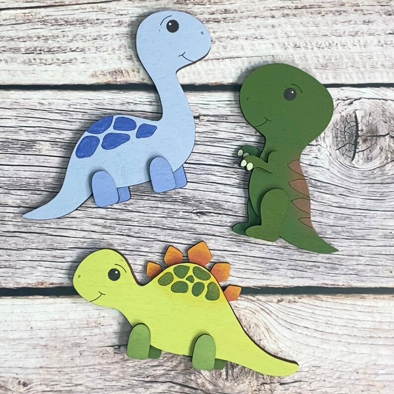 May include: Three wooden dinosaur figurines. A blue dinosaur with a light blue and dark blue patterned back, a green dinosaur with a brown stripe down its back, and a green and yellow dinosaur with a green and orange patterned back.