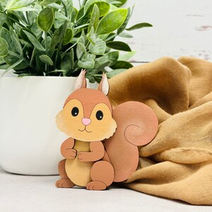 May include: A wooden squirrel figurine with a brown body, a white belly, and a fluffy tail. The squirrel is standing on a brown fabric surface.