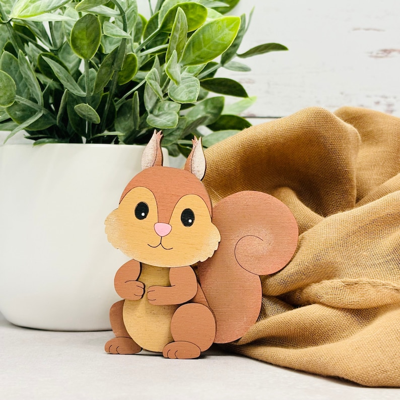 May include: A wooden squirrel figurine with a brown body, a white belly, and a fluffy tail. The squirrel is standing on a brown fabric surface.