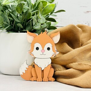 May include: A wooden cutout of a fox with a white chest and a fluffy tail. The fox is orange and has black eyes and a pink nose.
