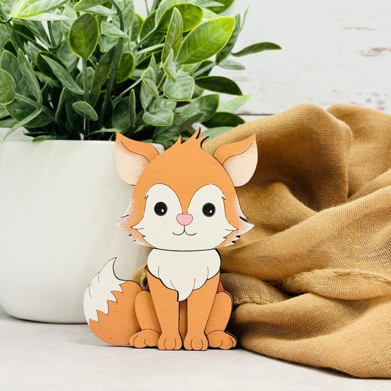 May include: A wooden cutout of a fox with a white chest and a fluffy tail. The fox is orange and has black eyes and a pink nose.