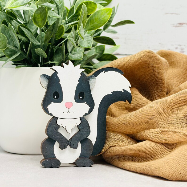 May include: A black and white wooden cutout of a skunk with a white face and black eyes. The skunk is standing on a brown fabric surface.