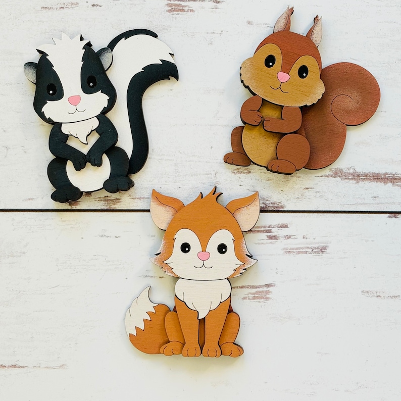 May include: Three wooden animal figurines: a black and white skunk, a brown squirrel, and a brown fox. The animals are all cartoon-like and have friendly faces.