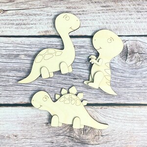 May include: Three wooden dinosaur shapes, ready to be painted. The dinosaurs are a stegosaurus, a brachiosaurus, and a tyrannosaurus rex.