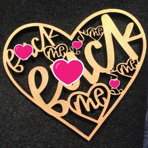 Puede incluir: Un letrero de madera en forma de corazón dorado con las palabras "Back Me Back Me Back Me" recortadas. El letrero tiene cinco corazones rosas.