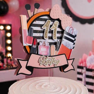 May include: A white frosted cake topped with a decorative topper. The topper features a black and white striped background with makeup and shopping bag illustrations. The number 11 is in gold glitter, and the name "Kathy" is displayed on a banner.
