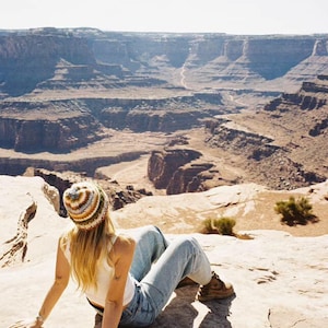 May include: A person wearing a striped beanie, white tank top, and blue jeans, sitting on a rock with a scenic canyon backdrop. The hat features cream, brown, and blue stripes. The landscape is a mix of tan and brown rock formations.
