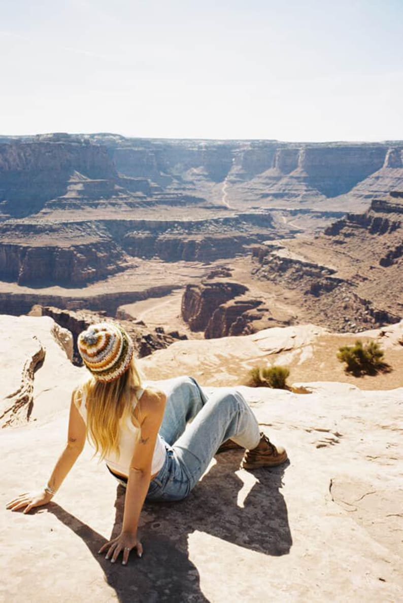 May include: A person wearing a striped beanie, white tank top, and blue jeans, sitting on a rock with a scenic canyon backdrop. The hat features cream, brown, and blue stripes. The landscape is a mix of tan and brown rock formations.