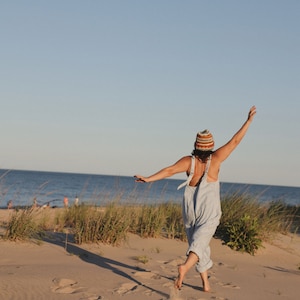 May include: A person wearing a striped knit hat and light blue overalls, standing on a sandy beach with arms outstretched. The background features the ocean and a clear blue sky, with tall grass and sand dunes.