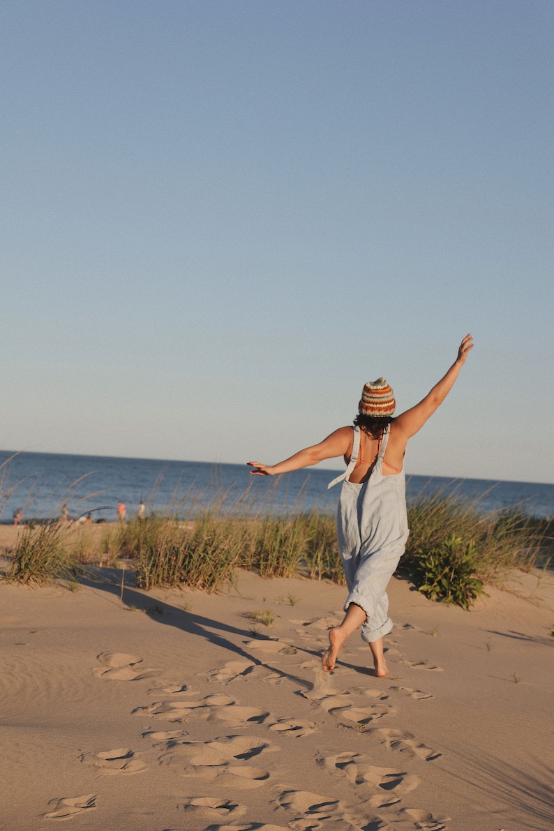May include: A person wearing a striped knit hat and light blue overalls, standing on a sandy beach with arms outstretched. The background features the ocean and a clear blue sky, with tall grass and sand dunes.