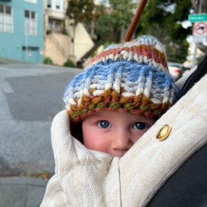May include: A baby wearing a knitted hat with blue, white, red, and brown stripes, peeking out from a beige baby carrier.