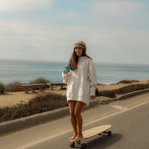 May include: A person wearing a white hoodie dress and a patterned beanie stands barefoot on a longboard, holding a teal mug. The scene is set on a coastal road with the ocean in the background under a blue sky.