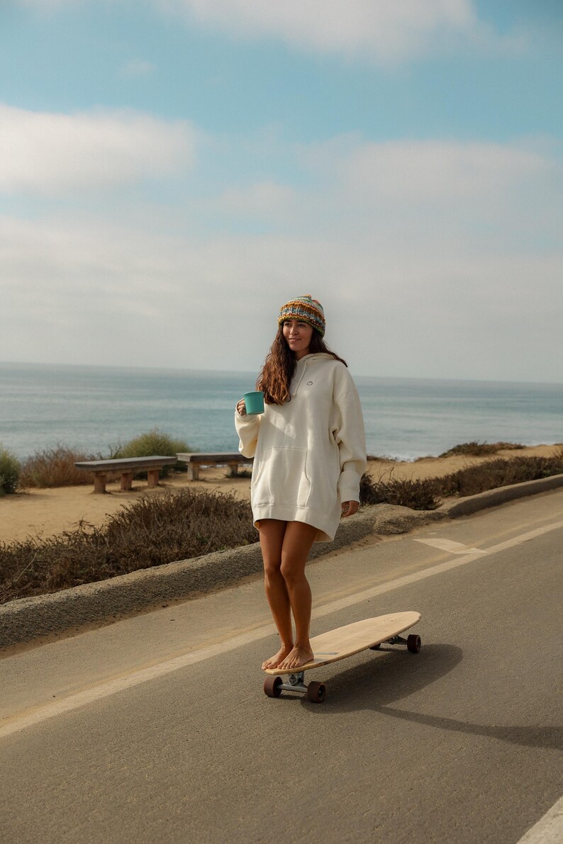 May include: A person wearing a white hoodie dress and a patterned beanie stands barefoot on a longboard, holding a teal mug. The scene is set on a coastal road with the ocean in the background under a blue sky.