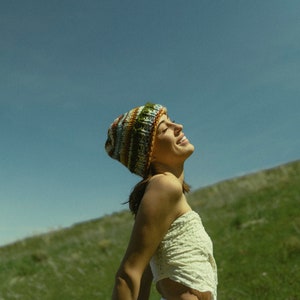 May include: A colorful knit beanie with stripes of green, brown, and blue. The beanie is worn with a cream-colored, strapless top and white shorts. The background features a green hillside and a clear blue sky.
