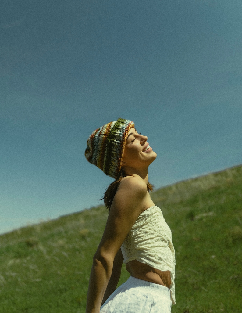 May include: A colorful knit beanie with stripes of green, brown, and blue. The beanie is worn with a cream-colored, strapless top and white shorts. The background features a green hillside and a clear blue sky.