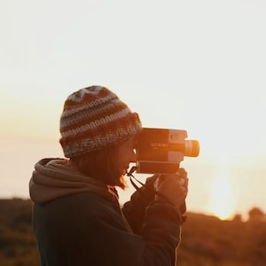 May include: A person wearing a knit hat and a hooded jacket is holding a vintage film camera, capturing a sunset. The camera is black and orange, with a lens and viewfinder. The background features a golden sky and a blurred landscape.
