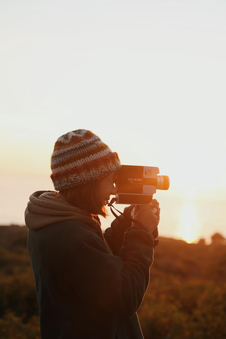 May include: A person wearing a knit hat and a hooded jacket is holding a vintage film camera, capturing a sunset. The camera is black and orange, with a lens and viewfinder. The background features a golden sky and a blurred landscape.
