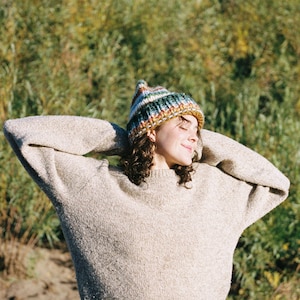 May include: A person wearing a beige sweater and a colorful knit beanie with stripes of green, blue, orange, and yellow. The person is smiling with their arms raised, and the background is blurred green foliage.