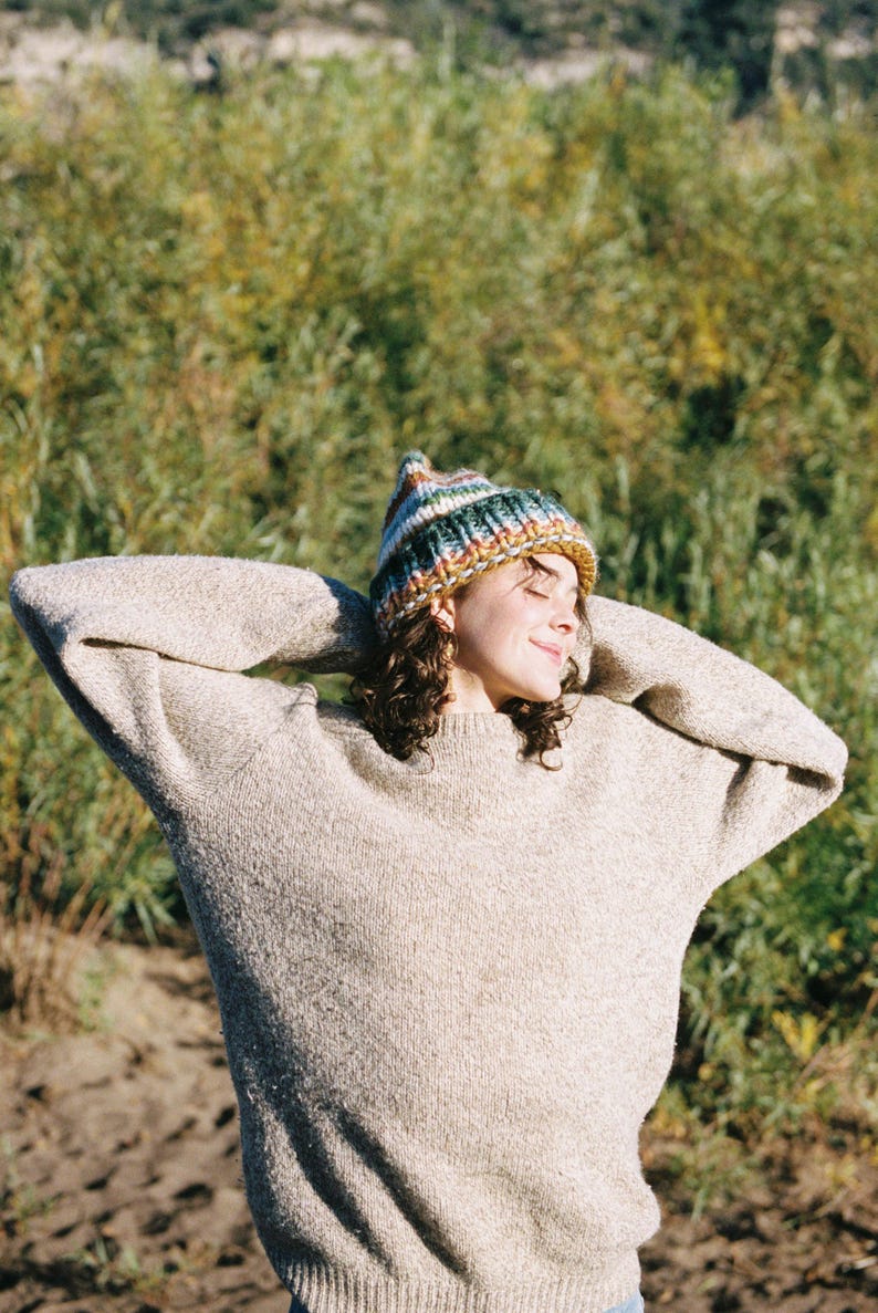 May include: A person wearing a beige sweater and a colorful knit beanie with stripes of green, blue, orange, and yellow. The person is smiling with their arms raised, and the background is blurred green foliage.