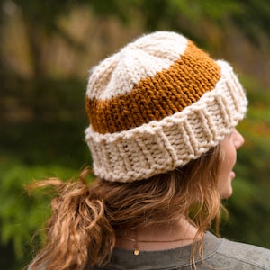 May include: A white and brown knitted beanie with a thick, chunky knit texture. The beanie has a wide brim and a curved shape.