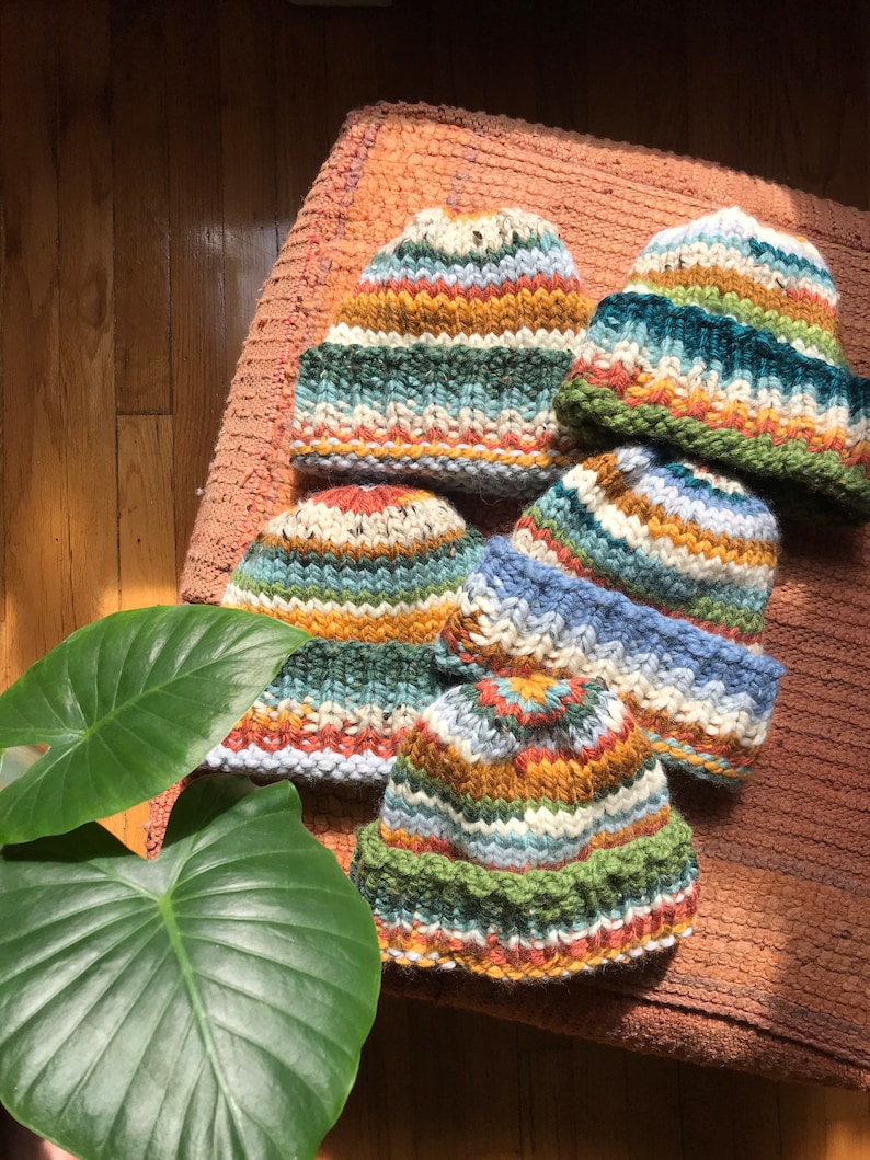 May include: A collection of hand-knitted beanies in various colors, including green, blue, orange, and white. The hats are arranged on a textured, peach-colored surface, with a large green leaf in the foreground. The beanies have a striped pattern.