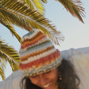 May include: A colorful, striped knit beanie in shades of red, orange, yellow, blue, and white. The hat is worn under a palm tree with green fronds against a bright blue sky. The beanie is a cozy accessory.
