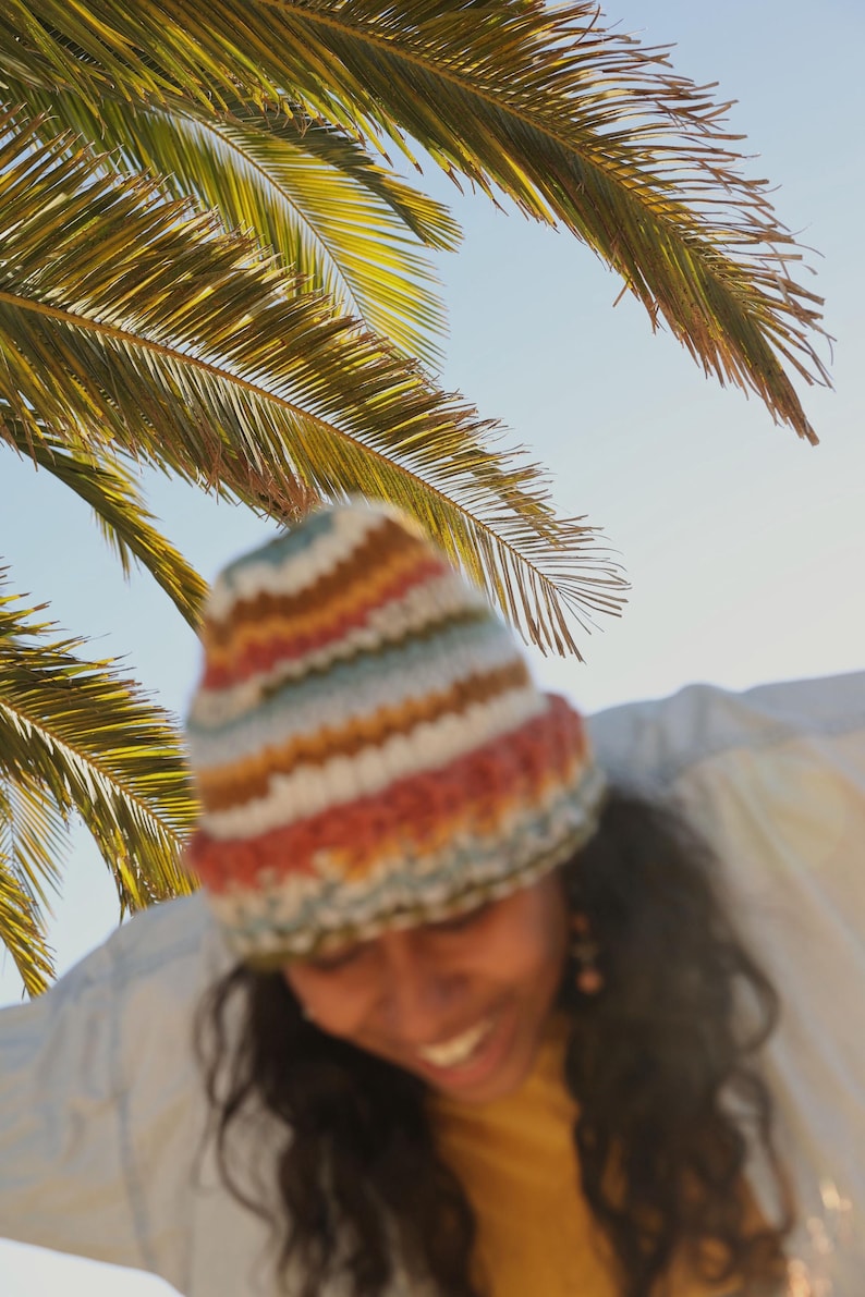 May include: A colorful, striped knit beanie in shades of red, orange, yellow, blue, and white. The hat is worn under a palm tree with green fronds against a bright blue sky. The beanie is a cozy accessory.