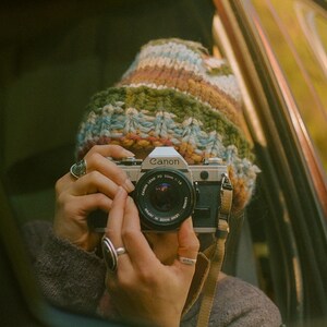 May include: A vintage Canon camera with a black lens is held by a person wearing a colorful knit hat and rings. The camera has a tan strap and is positioned to take a photo. The background is blurred, suggesting an outdoor setting.