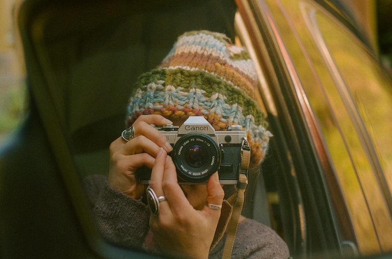 May include: A vintage Canon camera with a black lens is held by a person wearing a colorful knit hat and rings. The camera has a tan strap and is positioned to take a photo. The background is blurred, suggesting an outdoor setting.