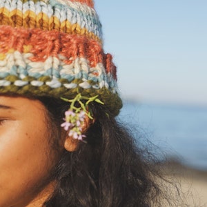 May include: A colorful, hand-knitted beanie in shades of orange, blue, green, and yellow. The hat is worn with long, dark hair and small pink flowers tucked behind the ear. The background is a clear blue sky and ocean.
