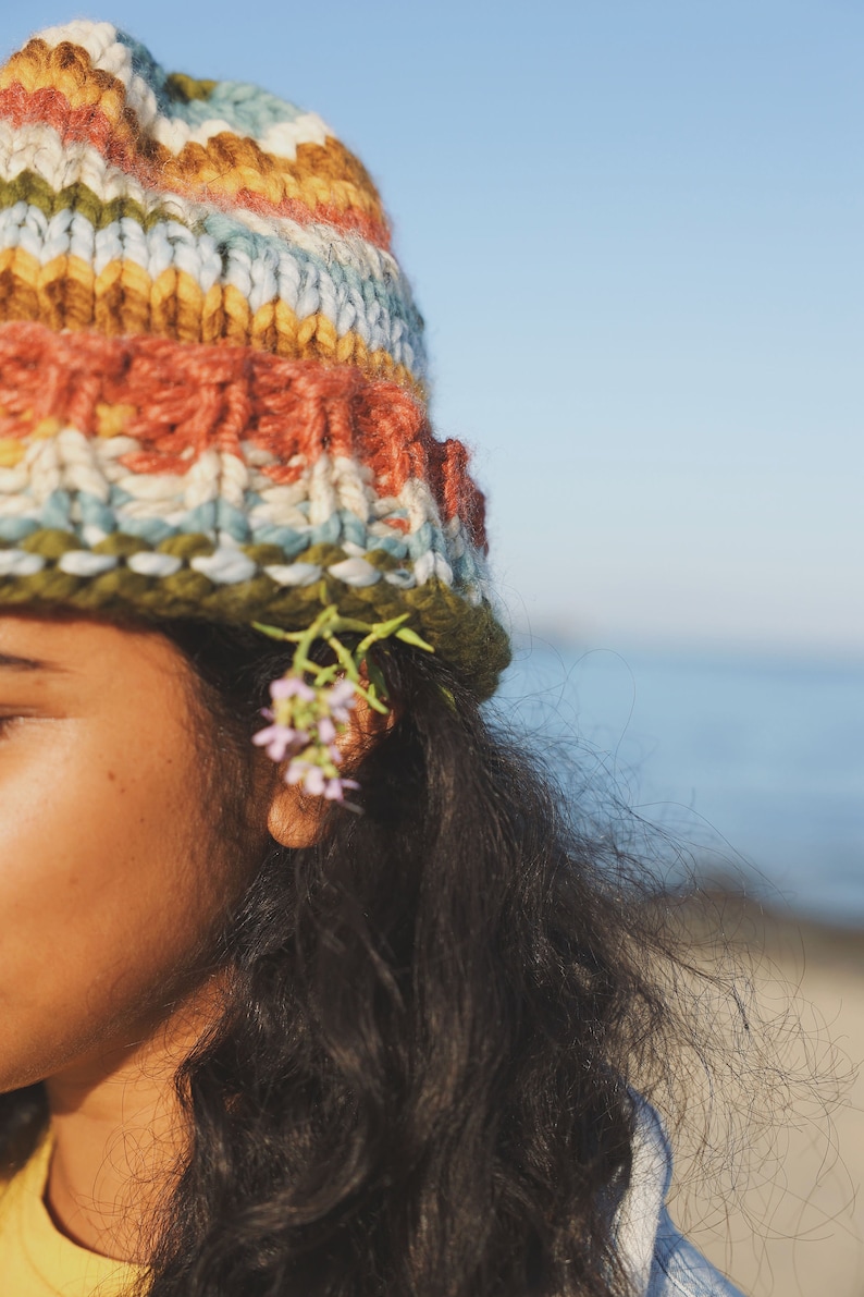 May include: A colorful, hand-knitted beanie in shades of orange, blue, green, and yellow. The hat is worn with long, dark hair and small pink flowers tucked behind the ear. The background is a clear blue sky and ocean.