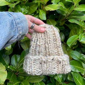 Może przedstawiać: Gruba wełniana czapka typu beanie w kolorze kremowym z brązowymi plamkami. Czapka jest wykonana z grubego włókna i ma żebrowany brzeg.