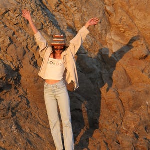 May include: A person wearing a white crop top with the text "OBOS", light wash jeans, a beige jacket, and a knit hat with sunglasses, standing in front of a rocky cliff. The person has their arms raised.