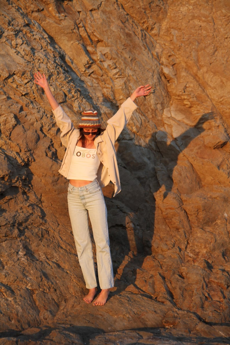May include: A person wearing a white crop top with the text "OBOS", light wash jeans, a beige jacket, and a knit hat with sunglasses, standing in front of a rocky cliff. The person has their arms raised.