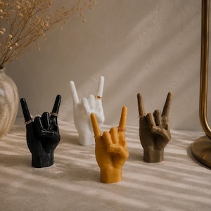Puede incluir: Cuatro esculturas con forma de mano en negro, blanco, amarillo y marrón, cada una mostrando un gesto de mano de rock and roll. Cada escultura presenta anillos finos de tono dorado en los dedos, sirviendo como soporte decorativo para joyas.