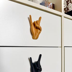 May include: Two 3D printed hand-shaped drawer pulls. One is gold and the other is black. Both hands are making the rock and roll hand gesture.