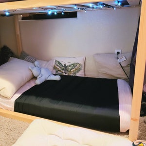 Puede incluir: Una almohada con forma de mariposa en blanco y negro se encuentra sobre una cama con una almohada blanca y un animal de peluche azul. La cama est&aacute; hecha con una s&aacute;bana negra y una manta blanca y rosa. Hay luces de cadena colgando de la litera superior de una litera.