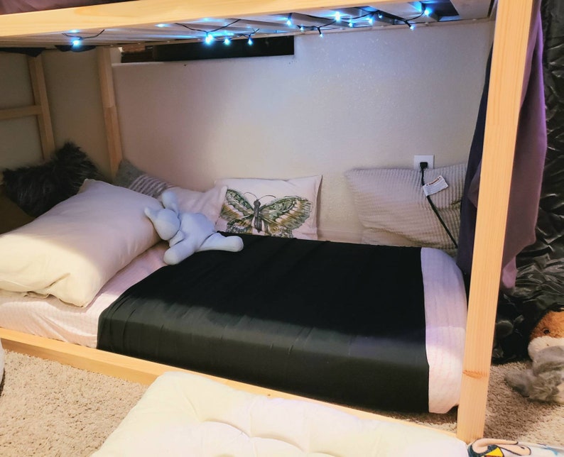 Puede incluir: Una almohada con forma de mariposa en blanco y negro se encuentra sobre una cama con una almohada blanca y un animal de peluche azul. La cama est&aacute; hecha con una s&aacute;bana negra y una manta blanca y rosa. Hay luces de cadena colgando de la litera superior de una litera.