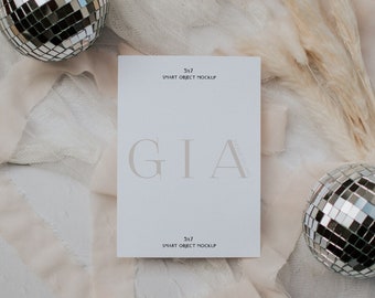 May include: A white card with the text "GIA" in a large, bold font. The card is surrounded by a white, sheer fabric and a light pink ribbon. There are two silver disco balls on either side of the card.